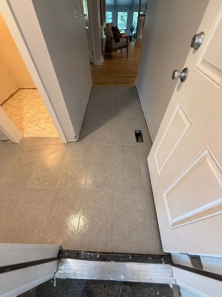 Grey speckled square floor tile in a hallway, with a rectangular floor register and a metal threshold at a wood floor